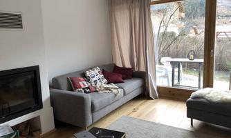 A modern living room with a gray sofa, several cushions, and a large window. Outside, there is a beautiful view of nature.