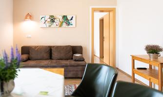 A cozy living room with a brown sofa and a table. A colorful picture hangs on the wall, and a table stands in the corner next to a plant.