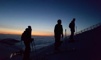 Three skiers stand silhouetted on a mountain as the sun rises. The sky is colored in soft shades of blue and orange.