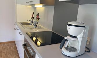 A modern kitchen with a stove, sink, and a coffee machine. The walls are light and the wooden floor creates a cozy atmosphere.