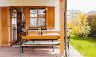 A cozy outdoor area with a bench and a table. The terrace has orange shutters and overlooks a green garden.