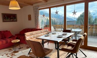 A cozy dining area with a large table and chairs. Large windows provide a beautiful view of the mountains and the balcony.