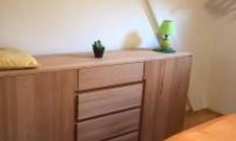 A cozy bedroom with a wood-colored wardrobe, a green lamp, and a small plant. The room has a bright, friendly atmosphere.