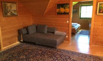 A cozy room with wooden paneling and a gray sofa. In the background, a bedroom with a window and carpet is visible.