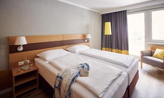 A modern hotel room with a double bed and an armchair. Large windows let in plenty of daylight.