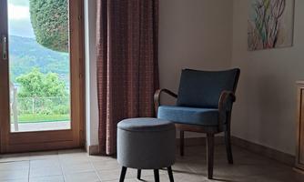 A cozy living space with a blue chair and a small table. Large windows provide a view of greenery and plenty of daylight.