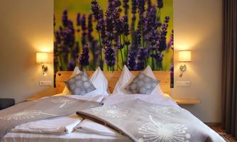 A cozy bedroom with a double bed and decorative pillows. In the background, there is a large wall mural of lavender.