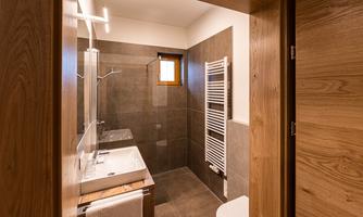 A modern bathroom with gray tiles and a sink. The shower is separated by a glass wall, and there is a towel radiator.