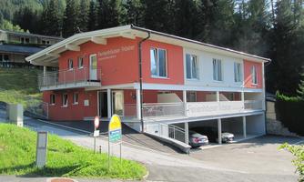 A modern holiday home with an orange paint job in a green environment. In the foreground, parked cars and trees are visible.