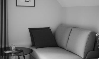 A cozy living area with a gray couch and a small table. A piece of artwork hangs on the wall, and the windows have dark curtains.