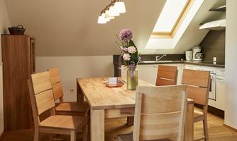 A bright dining room with a wooden table and four chairs. A vase with flowers is on the table, and the kitchen is well equipped.