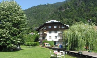 A beautiful house surrounded by green grass and tall trees. In the background, there are mountains and a clear sky.