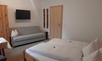 A cozy room with a double bed, a gray sofa, and a TV on the wall. The walls are painted in neutral colors and there are wood accents.