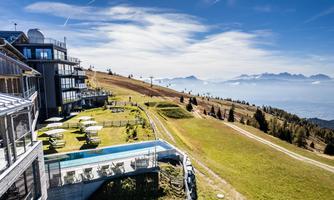 A modern hotel on a sunny mountain meadow with a view of the surrounding mountains. The pool area and loungers invite you to relax.
