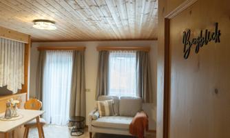A cozy living room with a bright sofa and a dining table. The windows are adorned with curtains, and the wooden ceiling gives a warm atmosphere.