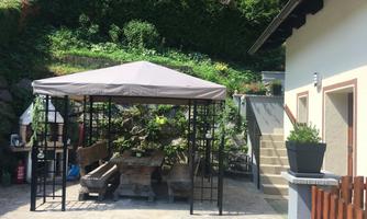 A cozy outdoor area with a pavilion and rustic seating. In the background, green plants and a part of the house are visible.