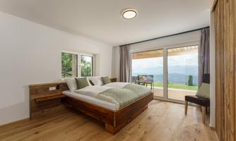A modern bedroom with a large bed and hardwood floor. The windows offer a beautiful view of the landscape.