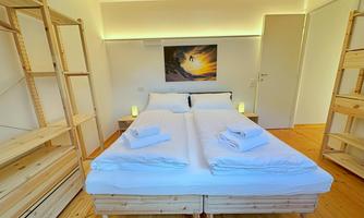 A cozy bedroom with a large bed and fresh towels. The decor is modern and minimalist with wooden shelves and a beautiful wall painting.