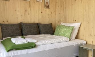 A cozy room with wooden paneling and a bed. On the bed are cushions and blankets in shades of green and gray.