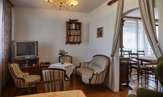A cozy living room with a table, armchairs, and a television. In the background, a dining area with chairs and a window is visible.