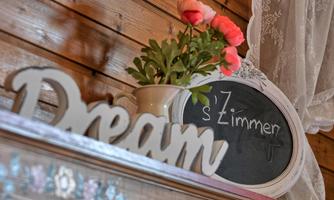 A decorative wooden shelf with the inscription "Dream" and a flower in a vase. In the background, there is a board with the writing "s'Zimmer".