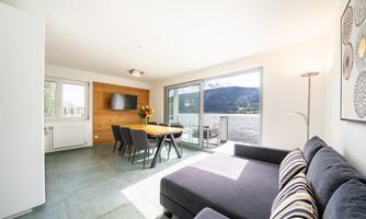 A bright and modern living space with a sofa and a dining table. Large windows offer a view of a mountainous landscape.