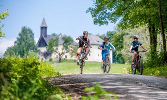 Three young cyclists ride on a forest path. In the background, a beautiful building and tall trees can be seen.