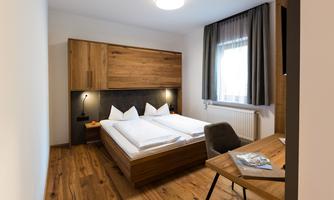 A modern bedroom with a wooden bed and two pillows. The room has large windows and a cozy atmosphere.