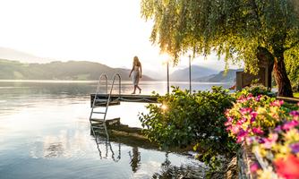 A woman is walking on a pier by the water. In the background, there are mountains and a blooming landscape.