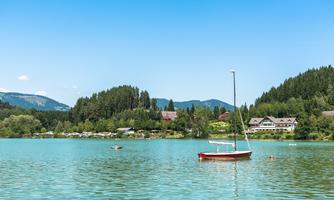 A tranquil lake with clear, turquoise water and a sailboat. In the background, green trees and vacation homes can be seen.
