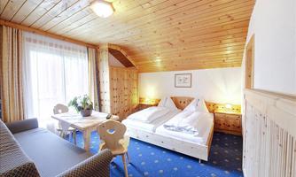 A cozy room with wooden paneling and a large window. It contains a double bed, a table, and an armchair.