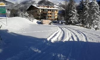 A snow-covered winter path leads to a cozy chalet. In the background, tall mountains are visible under a clear blue sky.