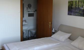 A simple bedroom with a double bed and two pillows. In the background, a bathroom is visible, and on the bed lies a small bouquet of flowers.