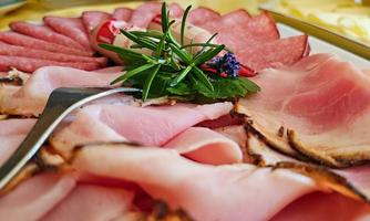 A selection of sliced sausages on a plate, decorated with fresh herbs. In the background, a basket of rolls can be seen.