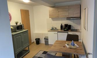 A modern kitchen unit with wooden cabinets and cooking utensils. A dining table with two chairs and a decorative element is visible in the foreground.
