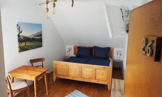 A cozy room with a wooden bed, blue pillows, and a small dining table. The walls are light, and there is a picture of a landscape on the wall.
