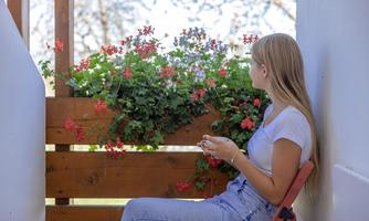 A young woman is sitting on a chair and looking at a flower balcony with red blossoms. Blurry trees are visible in the background.