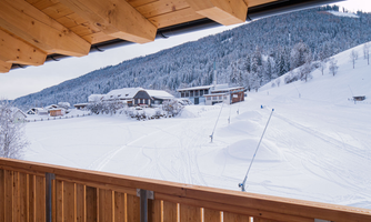 A peaceful winter view of snow-covered hills and a ski slope. In the background, some chalets and trees can be seen.
