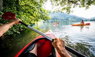 A person is paddling in a red kayak on calm water. In the background, other kayakers and green trees are visible.
