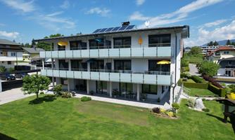 A modern residential building with three floors and a balcony. The green lawn and the blue sky give the surroundings an inviting atmosphere.