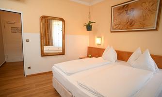 A bright room with a double bed and a large mirror. A decorative picture hangs on the wall, and there is a small plant hanger.