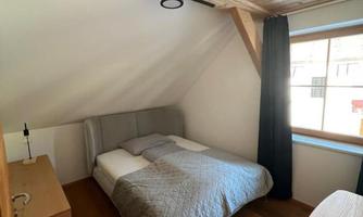 A cozy bedroom with a double bed and a wooden floor. The walls are light, and there is a window that lets in natural light.