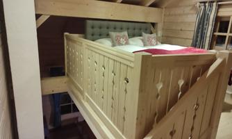 A cozy bedroom with a wooden bed on the upper floor. The walls and furniture are made of light wood, radiating a warm atmosphere.
