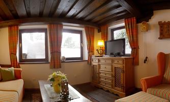 A cozy living room with wooden ceilings and large windows. There is a sofa, an armchair, and a dresser that create a warm atmosphere.