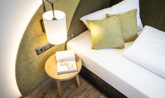 A modern bedroom with a bed, yellow cushions, and a small table. On the table, there are glasses and an open book.
