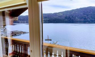 A view from a window overlooking a tranquil lake and surrounding hills. The balcony with a wooden railing offers a beautiful view of nature.