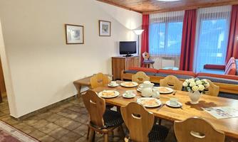 A cozy living space with a dining table and chairs. The room has large windows with curtains and a warm wooden ceiling.