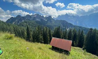 A green meadow with a small, red house and majestic mountains in the background. The sky is blue with some clouds.