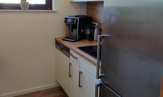 A modern kitchen with a refrigerator and a coffee machine on the countertop. The room has bright walls and large windows that let in daylight.