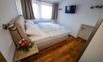 A cozy bedroom with a large double bed and neutral colors. The room has hardwood flooring and a window with curtains.
